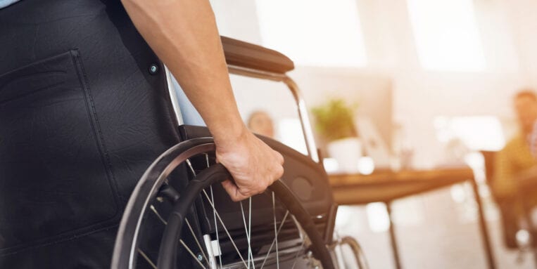A disabled man is sitting in a wheelchair. He holds his hands on the wheel. Nearby are his colleagues