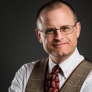 A headhsot of Dr.James. He is wearing glasses, a white dress shirt, red tie, and checkered brown vest. His hair is cut short and he smiling with no teeth at the camera