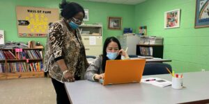 Linda Smith stand next to a student at a long table during a literacy class session.