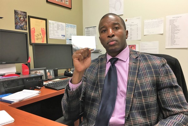 Ahmeed Turner sits behind a desk in a plaid suit, pink shirt, and tie