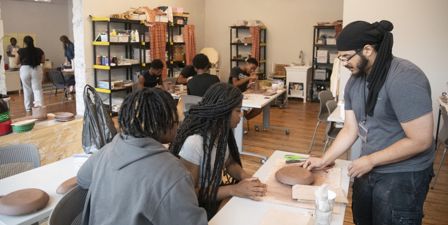 Person standing at table, with two people sitting at table with clay art